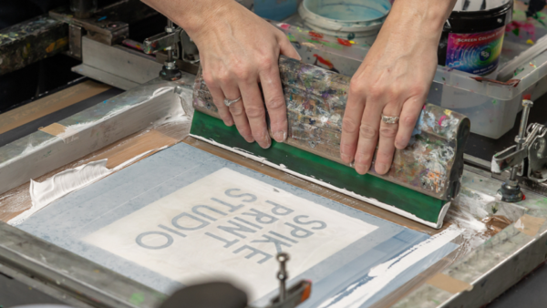 two hands pulling squeegee on screenprint frame to reveal text saying spike print studio